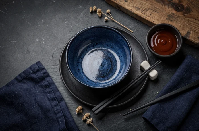 Flatlay de vaisselle artisanale en céramique bleu marine et noire mate sur ardoise sombre, avec baguettes et bol à tremper dans un style japonisant épuré
